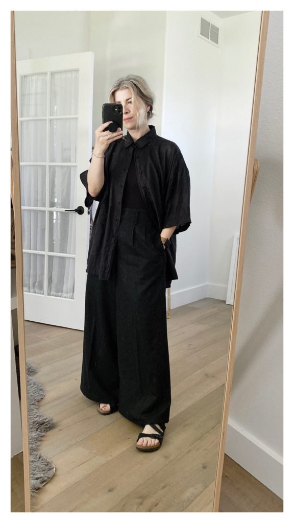 A mirror selfie of a small white woman with shoulder length blond hair that is pulled back in a ponytail. She is wearing an oversized black silk shirt over a black bodysuit and charcoal grey wide leg pants with black sandals. She has her right hand in her pocket.