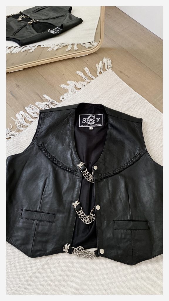  black leather vest is laid flat on a cream colored rug on a light wood floor. It is in front of a mirror and you ca see the reflection of the vest on the floor. The floor has leather braiding and chain closures with eagles on them.