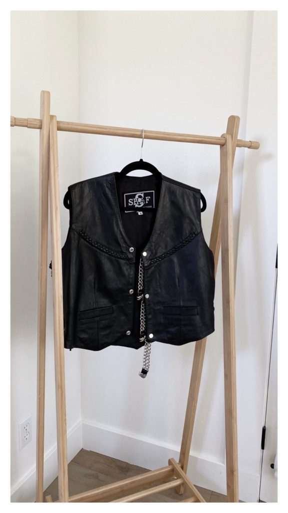 A black leather vest is hanging, alone, to dry from a light wood clothing rack.