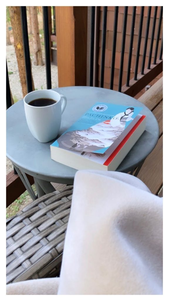 A table on a patio which is holding a cup of coffee in a white mug and the book, "Panchinko".