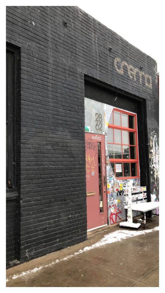 An image of a coffee shop in Denver Colorado called Cream Coffee House. The building is black brick and the name of the shop is painted in tan lettering. The door and window frame are red.