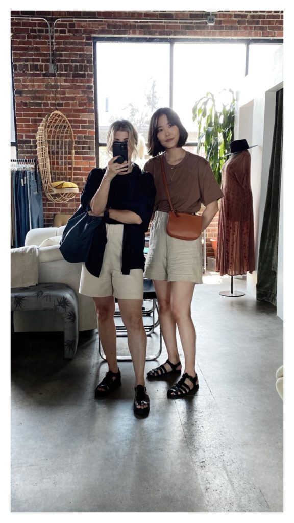 A mirror selfie of 2 women, one with blonde hair and one with brown. Both are wearing light colored shorts and fisherman sandals. The blonde has a black tank under a black short sleeved shirt that has been left open. The brunette has on a brown shirt with a light brown crossbody bag.