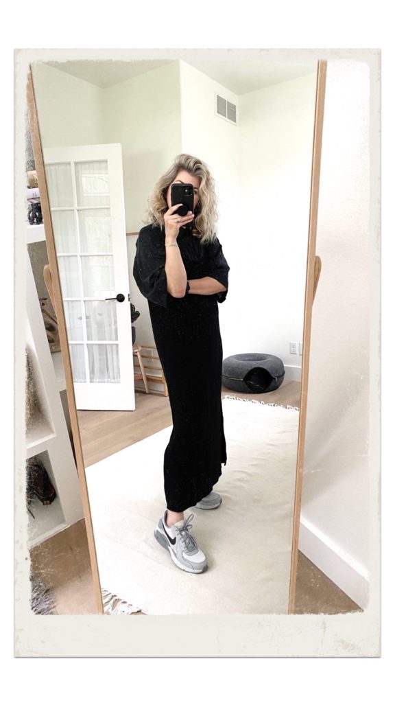 A mirror selfie of a small white woman who is wearing a short sleeved black silk short under a long black slip dress. She is wearing grey Nike sneakers.
