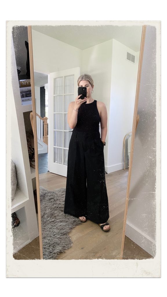 A mirror selfie of a small white woman who is wearing a black bodysuit with a pair of charcoal grey wide legged pants with flat black sandals. 