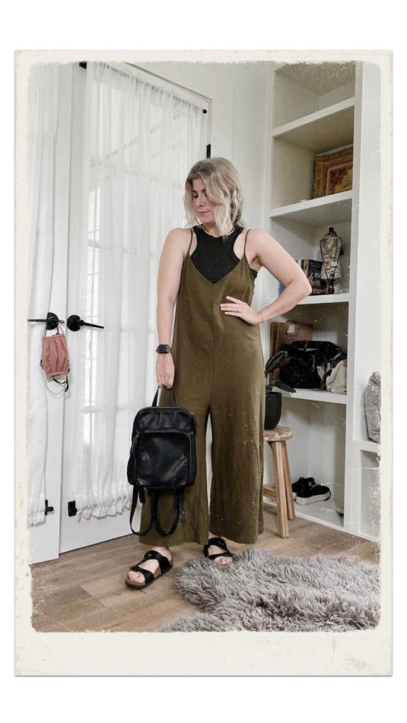 A small white woman is wearing an dark green tank top under an olive green jumpsuit with flat black sandals and she is holding a black backpack. she is standing own front of a built in bookshelf.