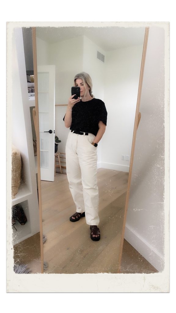 A mirror selfie of a small white woman who is wearing black t-shirt tucked into white full length.ants. the pant have a built in black belt. she is wearing brown fisherman sandals  