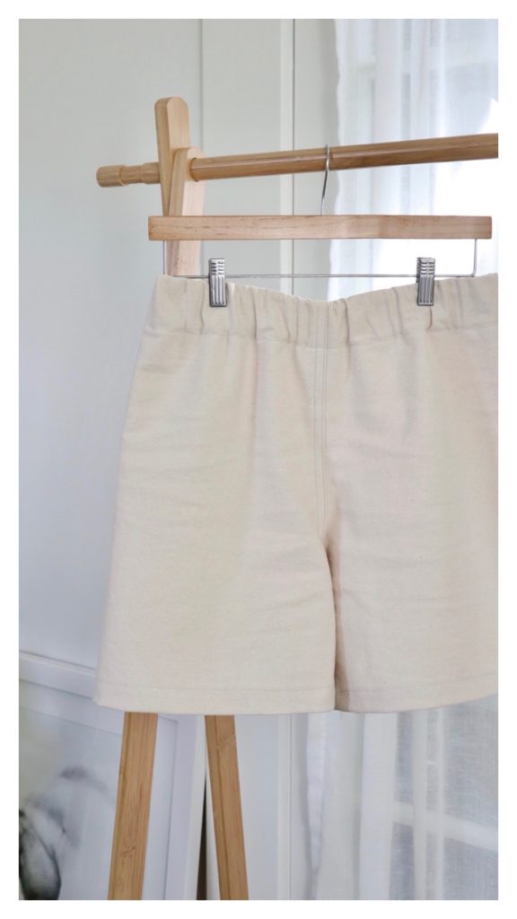 A pair of cream colored Pomona shorts are clipped to a light wood hanger and hanging from a light wood clothing rack.