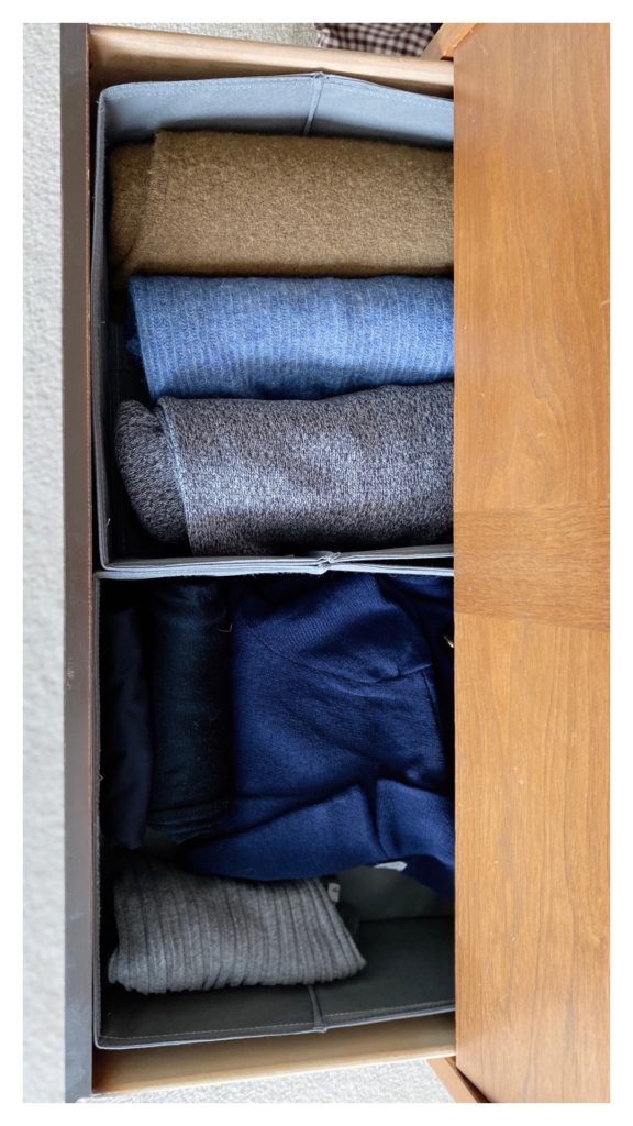 A view from above of the dresser drawer that holds a few of the capsule wardrobe items. There are 3 rolled sweaters in one basket: a brown, blue and marled grey/black one. In the basket beside it is a folded navy blue cardigan, a grey top and some black tops.