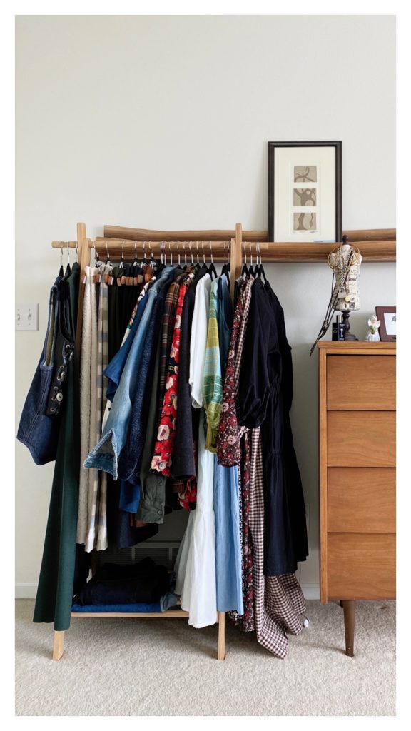 A wooden clothing rack is holding a selection of colorful clothing. There is a dresser to the right of it. You can partially see the dresser and a few items on it. There is a wooden picture rail above both items and one picture is in the middle.