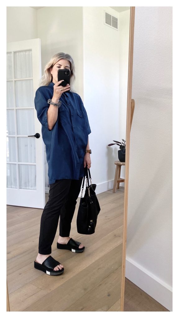 A mirror selfie of a small white woman who is turned slightly side on to the mirror. She is wearing a vibrant blue long silk shirt with shot sleeves over a pair of black straight leg pants with black leather slides. She is holding a black tote bag.