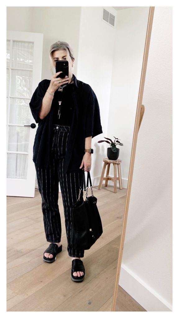 Mirror selfie of a small blonde woman who is wearing a black silk shirt over a black top and black jeans with white longitudinal stripes. She is holding a black tote bag in her right hadn't and wearing black leather slides.