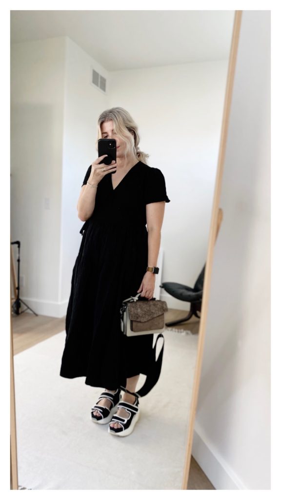 Mirror selfie of a small blonde woman who is wearing a long black wrap dress with short sleeves and tiers with a pair of black and white platform sandals and a grey satchel handbag.