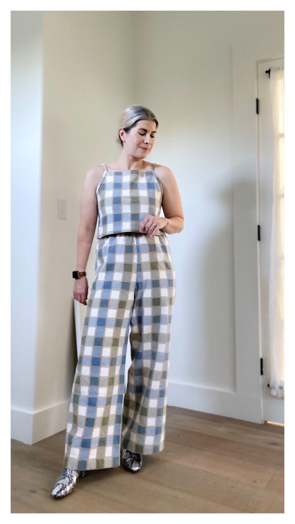 A small blonde woman is standing in front of a white wall and wearing a blue/green/white checkered cami with a pair of matching wide legged pants. She is wearing silver snakeskin boots.