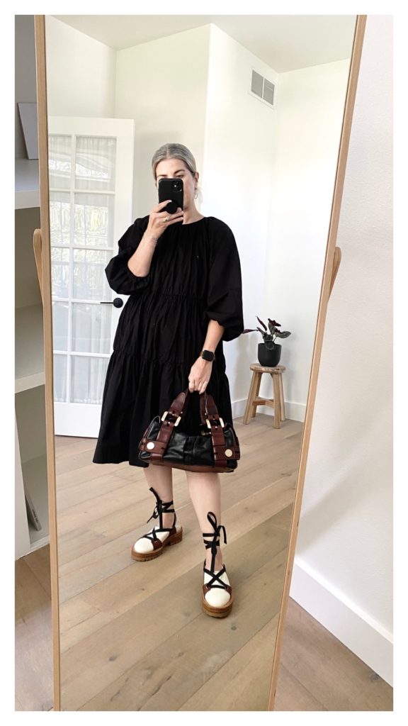 Mirror selfie of a small blonde woman who is wearing a long sleeved black tiered dress that ends just below her knees. She is wearing it with a pair of cream shoes that have 2 brown tabs on each side of each shoe. a black lace goes through the tabs and criss crosses over the shoe and then laces around the leg before being tied into a bow. She is holding a black leather satchel with brown handles and gold hardware.