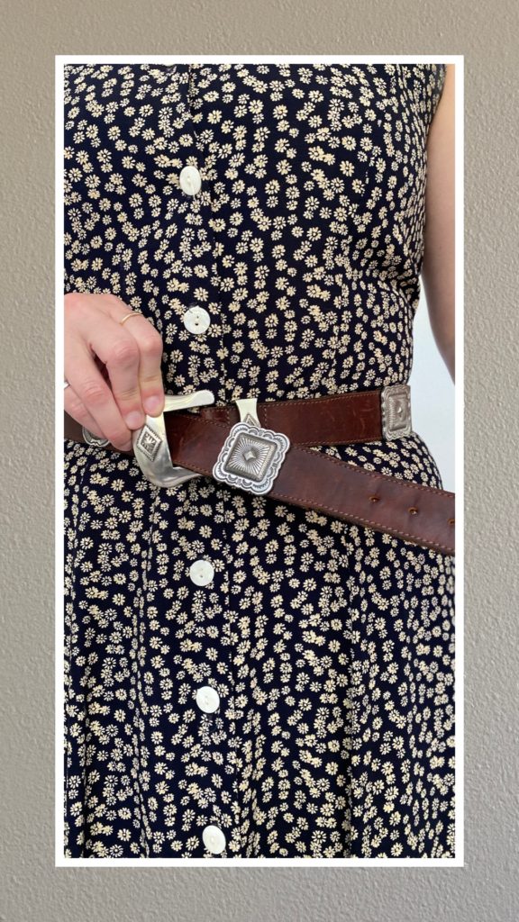 A partial photo of a woman's torso. She is wearing a navy dress with small tan flowers all over it. She has a brown leather belt around her waist and is holding it as tight as it can go. There are silver squares all around it at equal distances. It has a silver buckle.