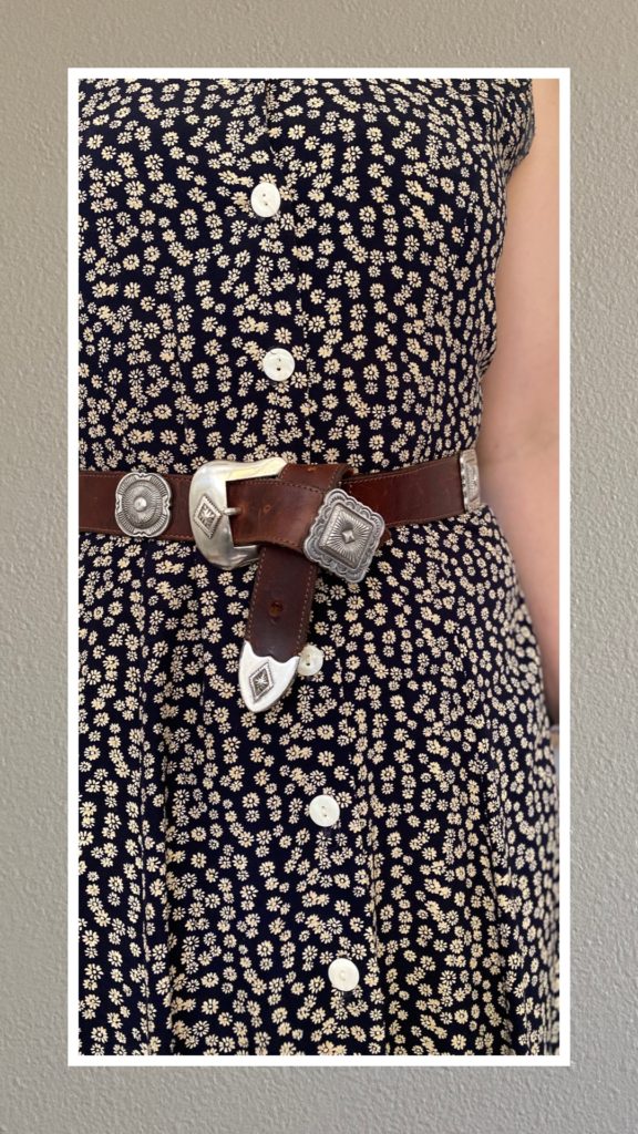 A partial photo of a woman's torso. She is wearing a navy dress with small tan flowers all over it. She has a brown leather belt around her waist that has been fixed to be able to secure it with the buckle. She has also twisted the end that was hanging down into a knot next to the silver buckle. There are silver squares all around it at equal distances.