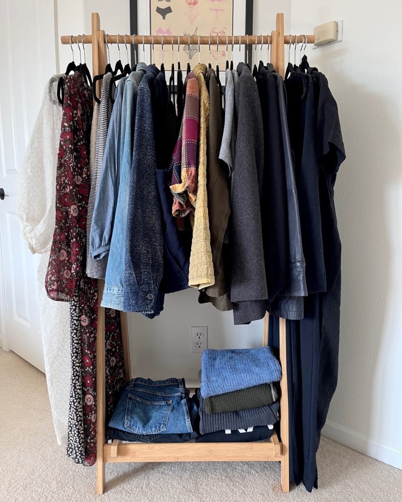 My Spring Capsule Wardrobe. 33 clothing items are included on a light wooden storage rail. items such as dresses, shirts, tops, pants and jackets are hanging on the rail while heavier items such as jeans and sweaters are folded and placed on the wooden rack at the bottom.