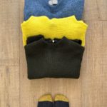 3 sweaters, which will be a part of my spring capsule wardrobe, partially stacked on top of each other, lay on a light wood floor. A medium blue sweater is at the top of the photo, slightly covered by a bright yellow sweater which is partly covered by an olive green sweater. Part of a pair of feet wearing mustard yellow/charcoal grey and light grey socks are visible at the bottom of the photo.