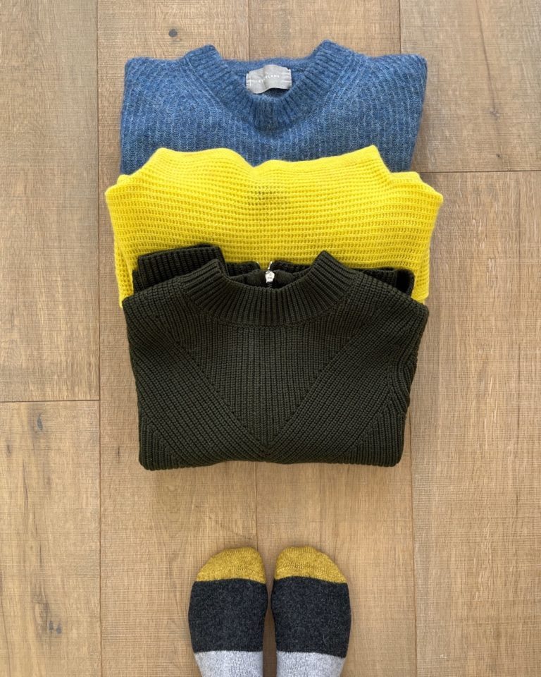 3 sweaters, which will be a part of my spring capsule wardrobe, partially stacked on top of each other, lay on a light wood floor. A medium blue sweater is at the top of the photo, slightly covered by a bright yellow sweater which is partly covered by an olive green sweater. Part of a pair of feet wearing mustard yellow/charcoal grey and light grey socks are visible at the bottom of the photo.
