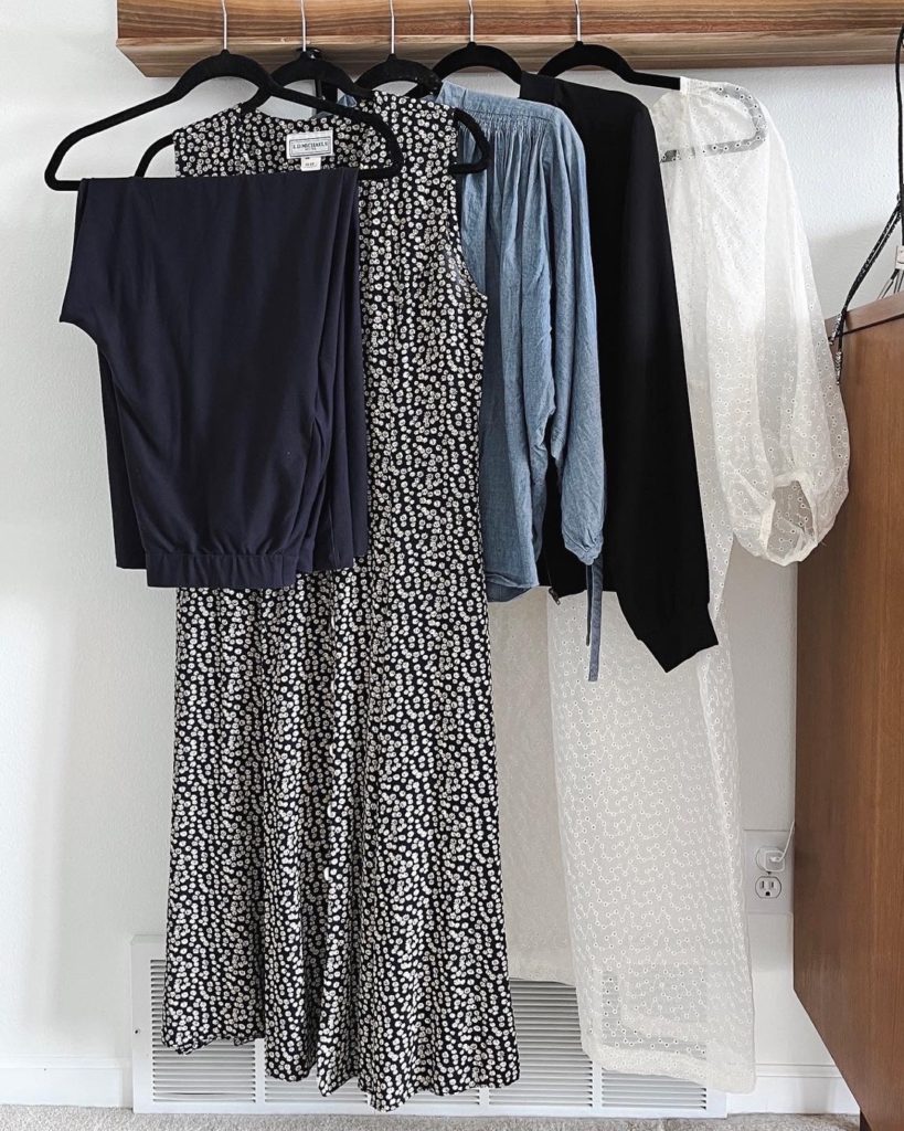 The 5 items in my mini capsule wardrobe are hanging from a wooden picture rail and against a white wall. From left to right, they are a pair of navy wide leg pants, a sleeveless navy dress with a button front and small tan flowers all over, a chambray blue blouse, a black silk jacket and a long white sheer dress with voluminous sleeves.