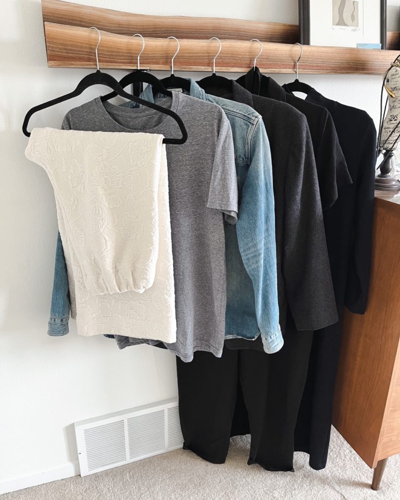 6 items that I removed from my spring capsule wardrobe hang from a wooden picture rail. From left to right: a pair of cream quilted pants, a grey t-shirt, a light wash denim shirt, a charcoal grey wool blazer, a short sleeve black jumpsuit, and a long mockneck black dress.