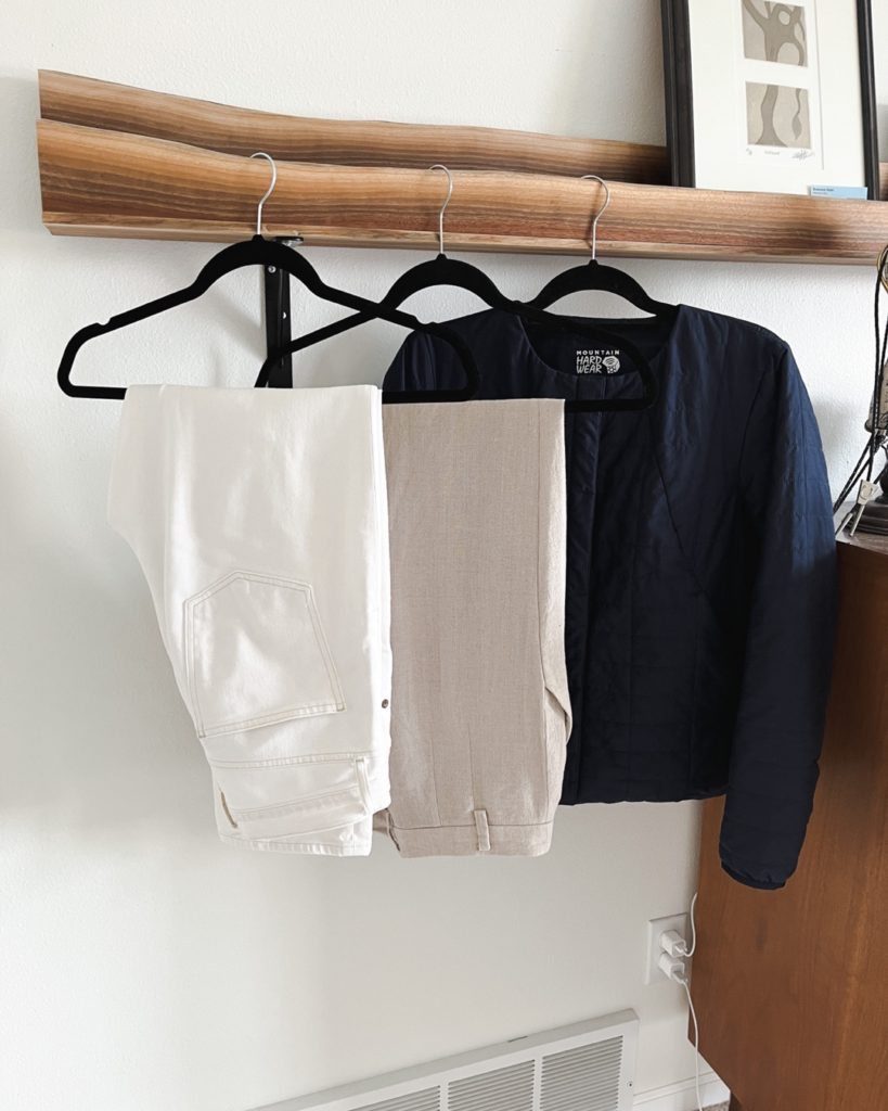 3 items that I added to my spring capsule wardrobe hang from a wooden picture rail. From left to right: a pair of white jeans, a pair of tan pants and the navy quilted jacket.