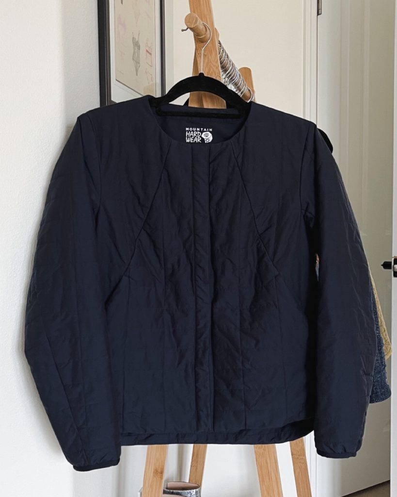 A navy quilted jacket with no collar and a rounded hem is hanging on the end of a wooded clothing rack.