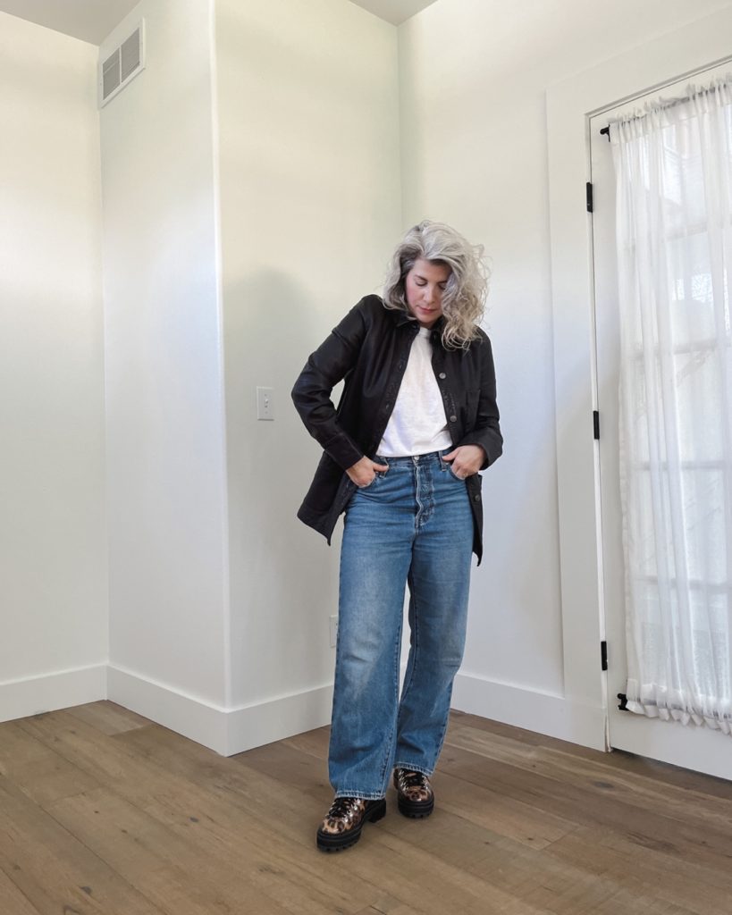 A petite white woman with silver curly hair is standing in front of a white wall. she is wearing a black leather jacket over a white top and baggy blue jeans. She is wearing leopard print hiking shoes.