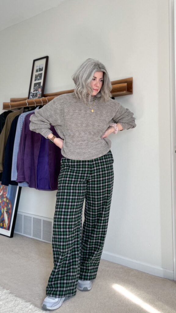 Wearing my first make nine sewing project. A small white woman with silver shoulder length hair is standing with her hands on her waist. She is wearing a grey textured sweater with the pants and grey sneakers.