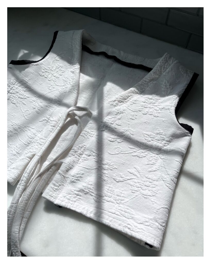 A white vest with a raised floral pattern is laid one top of a white surface. it has 4 front strings (2 on each side) and the armholes have black binding all the way around. The shadow of a windowpane is visible.