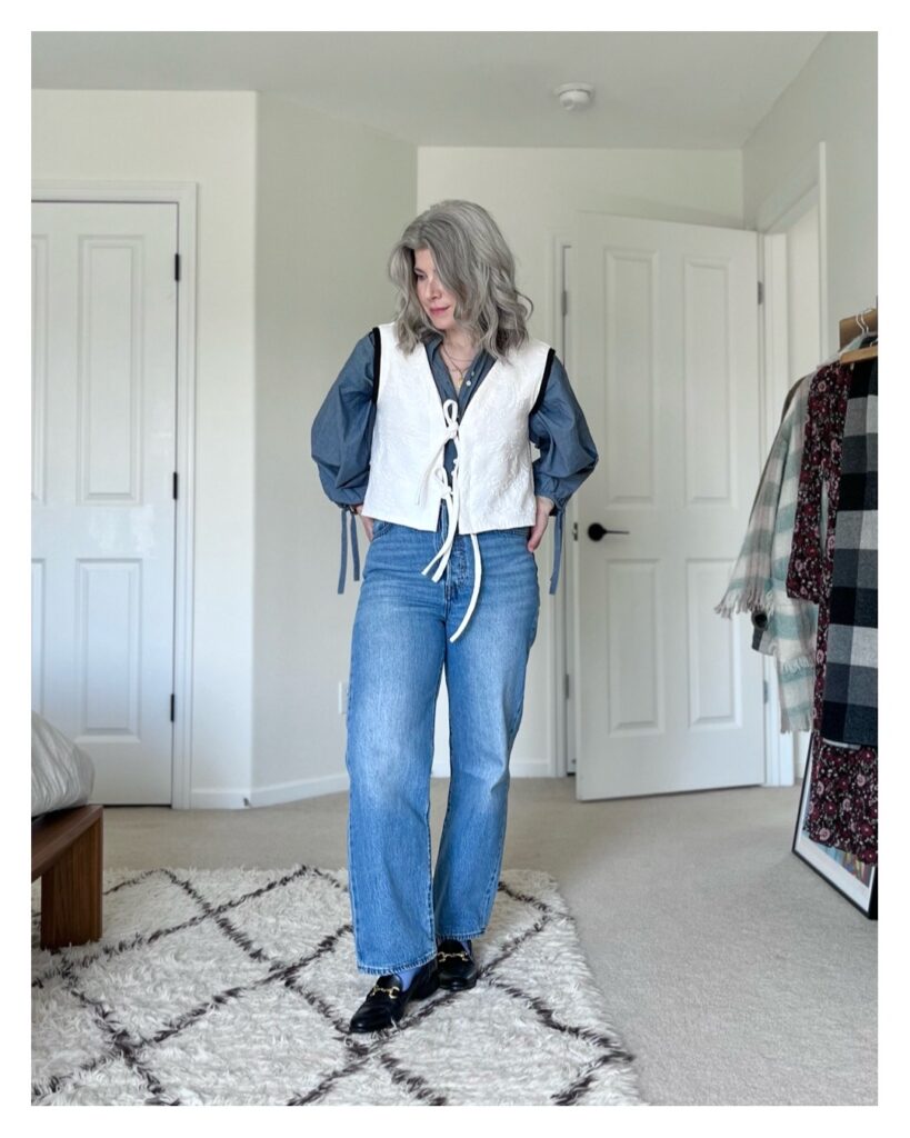 A small white woman with silver wavy hair is wearing the white vest (floral side out) over a blue blouse. The blouse has billowy sleeves and ties at the cuff. She is wearing straight leg blue jeans and black loafers with a brass hardware detail across the top.