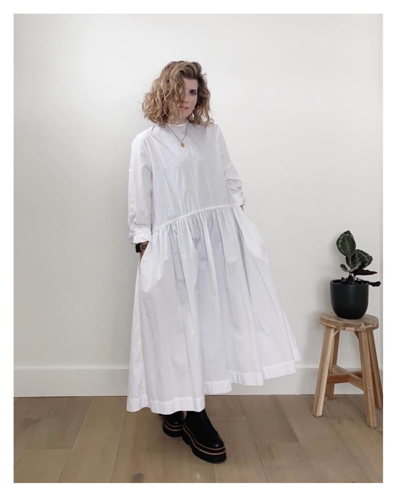 The original dress shortly after I received it. The photo is of a small white woman with blonde shoulder length curly hair. She is wearing a maxi length white dress with long sleeves and a dropped waist. She is wearing it with black platform boots.