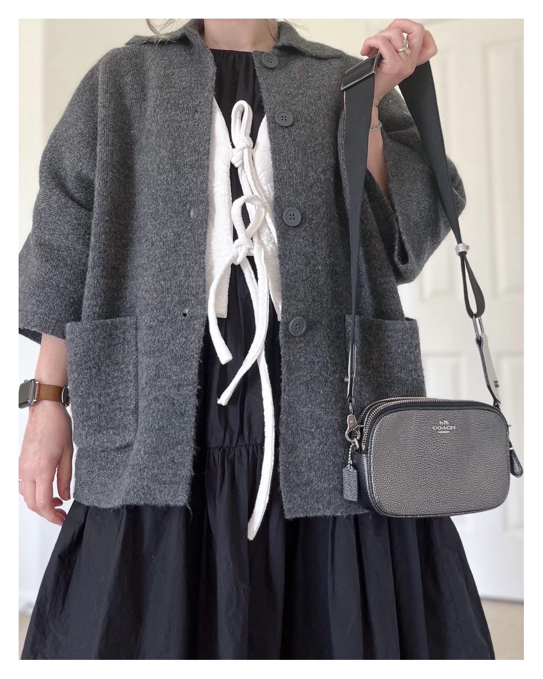 A small white woman is wearing the cardigan before the upcycle over a white vest with front ties and a midi length black dress. She is holding a silver handbag. The photo is from the woman's shoulders to her knees.