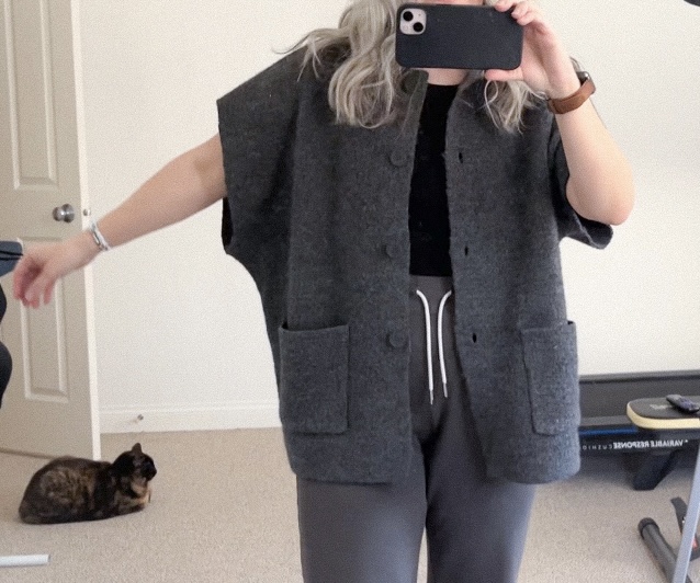 During the upcycle project...asmall white woman takes a mirror selfie with her left arm extended to show the cardigan sleeve after the first cut which removed the sleeve cuff.