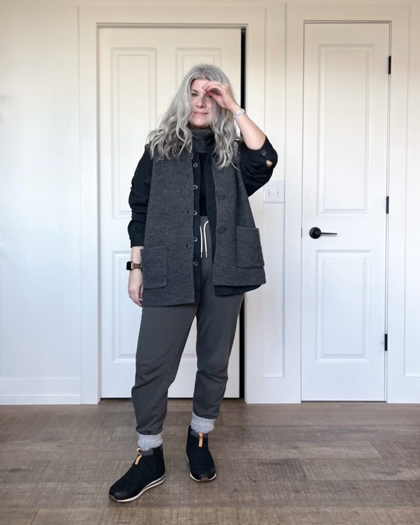 A small white woman is standing in front of a white wall and 2 white doors. She is wearing the charcoal grey vest over a black chore shirt and black tank top, with charcoal grey sweatpants that are tucked into light grey socks with black quilted boots.